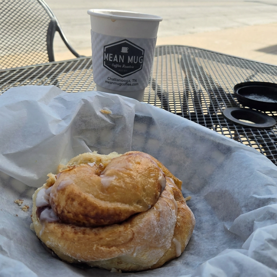 Breakfast sandwich on a bagel sits in paper wrapping on an outdoor metal café table beside a Mean Mug coffee cup.