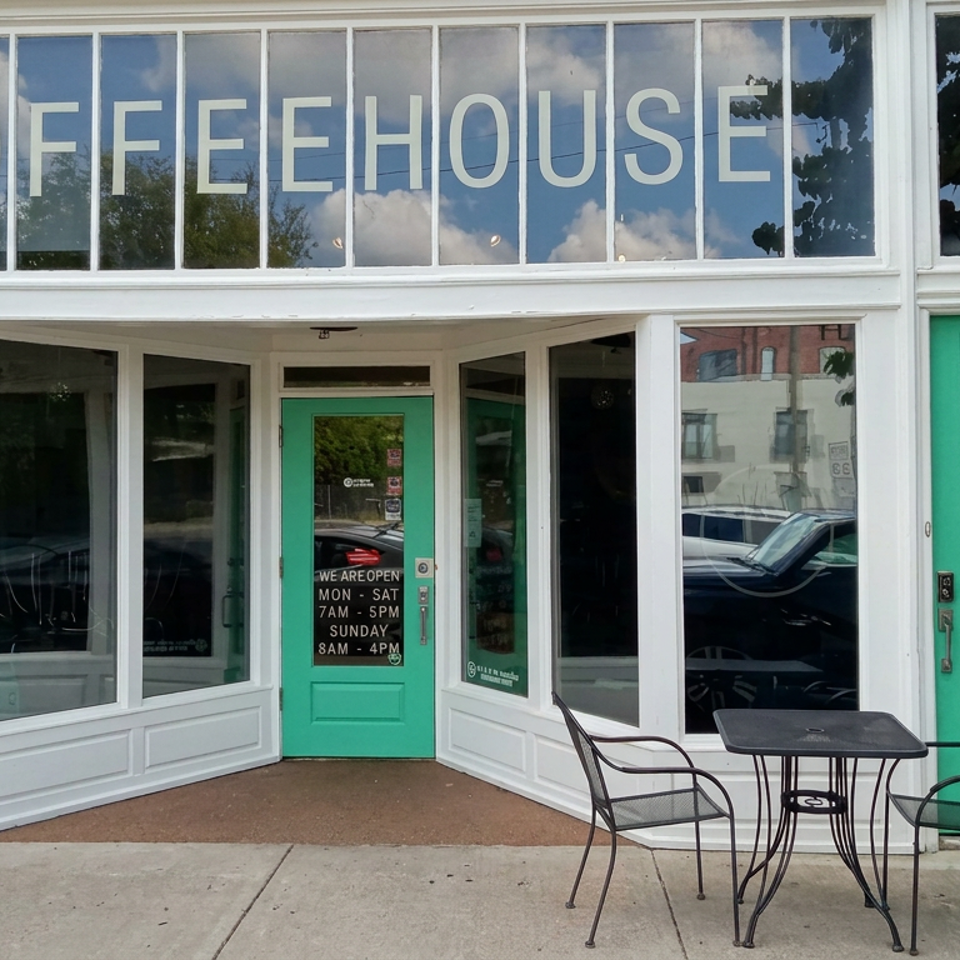 Coffeehouse storefront with large window lettering, turquoise doors, brick walls, and outdoor café tables and chairs.
