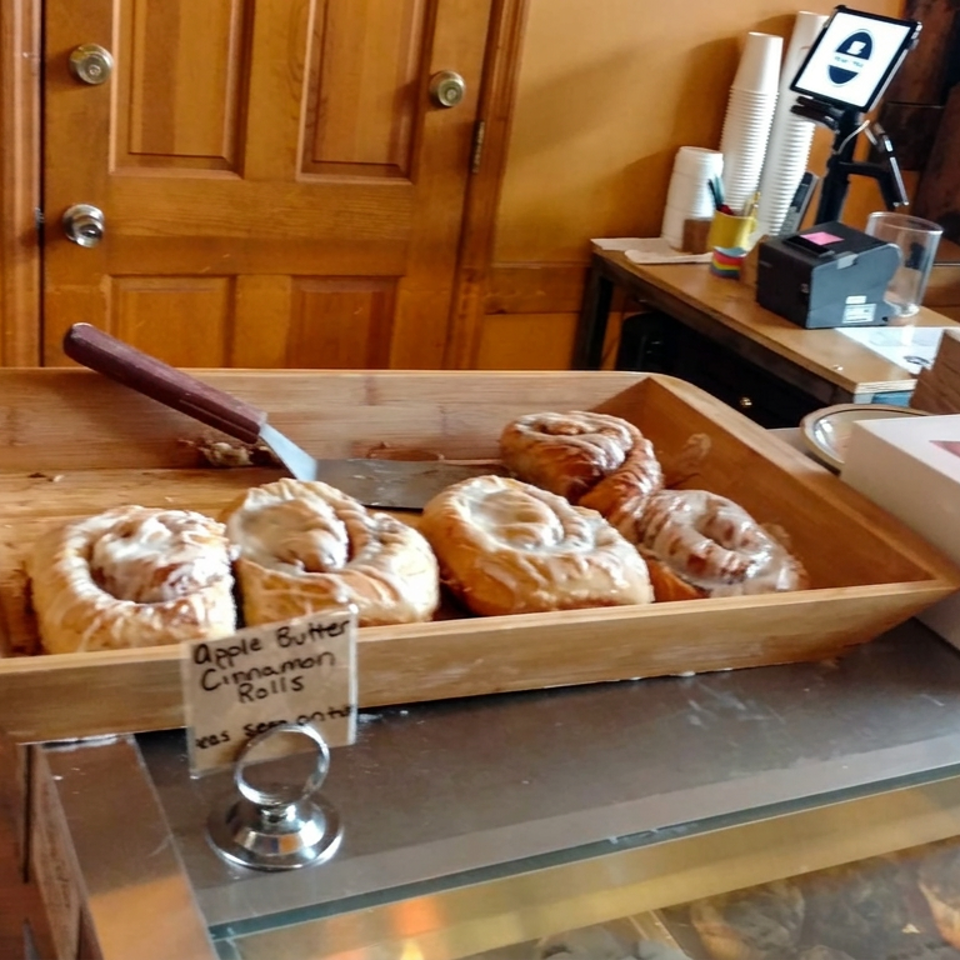 Wooden bakery display tray holding iced apple butter cinnamon rolls beside a boxed pastry on a café counter.